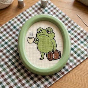 May include: An oval-shaped light green wooden frame containing a cross-stitch embroidery on white fabric. The design features a green frog with a tired expression holding a small white coffee cup in one hand and a brown suitcase in the other.