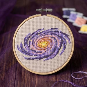May include: A circular wooden embroidery hoop containing a cross-stitch pattern of a spiral galaxy. The galaxy features a bright yellow centre transitioning into shades of orange, pink, and deep purple, with small white stitches representing stars.