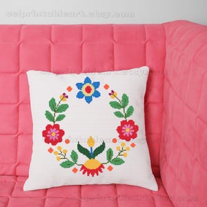 May include: White square pillow with a cross-stitch floral wreath design. The wreath features red, blue, yellow, and green flowers and leaves. The pillow is on a pink couch.