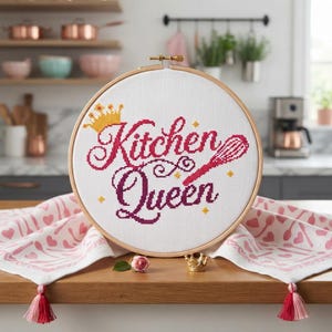 May include: A circular wooden embroidery hoop containing white fabric with cross-stitched text that reads Kitchen Queen in dark pink, accompanied by a yellow crown icon and a pink whisk icon, resting on a white cloth with pink kitchen patterns.