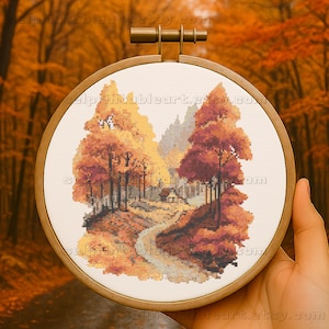 Può includere: Un paesaggio a punto croce di una foresta autunnale con alberi nei toni dell'arancione e del rosso. Un sentiero tortuoso conduce a una piccola casa. L'opera d'arte è esposta in un telaio da ricamo rotondo in legno.