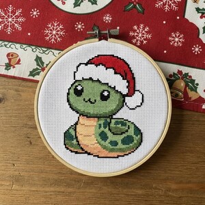 Op de afbeelding: Een ronde houten borduurring met een kruissteekpatroon van een groene slang met een lichtbruine buik. De slang heeft grote zwarte ogen en draagt een rood-witte kerstmuts op een witte stoffen achtergrond.