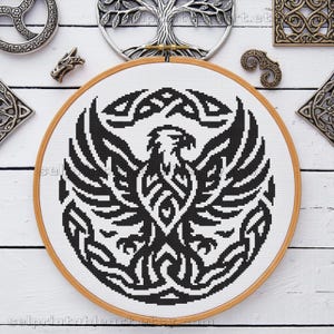 May include: A black and white cross-stitch design of a stylized eagle with outstretched wings, set within a wooden embroidery hoop. The design features intricate Celtic knotwork. Decorative metal pieces surround the hoop.