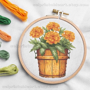 Puede incluir: Bordado de punto de cruz de flores de caléndula naranja en un cubo de madera, en un aro de madera. Hebras de hilo de bordar naranja, verde y amarillo están en el fondo. El texto "selprintableart.etsy.com" es visible.