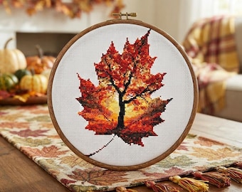 May include: A circular wooden embroidery hoop containing a cross-stitch pattern of a single large maple leaf. The leaf features a gradient of vibrant autumn colors including deep red, orange, and yellow, with a dark brown stem and veins.