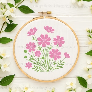 Può includere: Un telaio da ricamo a punto croce con un motivo floreale di fiori di cosmo rosa e foglie verdi. Il ricamo è su uno sfondo di tessuto bianco, incorniciato da un telaio di legno. Il testo "selprintableart.etsy.com" è visibile.