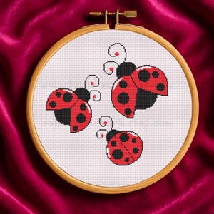 May include: A cross-stitch embroidery hoop featuring three ladybugs with red wings and black spots. The ladybugs are set against a white background, framed by a wooden hoop. The design is simple and charming.