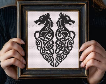 May include: Framed cross-stitch art featuring a symmetrical design of two black dragon-like creatures facing each other. The dragons are intertwined with intricate knotwork, set against a light pink background within a black frame.