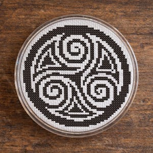 May include: A round, cross-stitched coaster featuring a black and white Celtic Triskele design. The coaster is encased in a clear plastic holder, set against a wooden background. The design showcases three interlocking spirals.