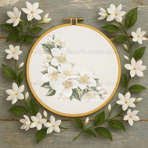 May include: A cross-stitch embroidery hoop featuring a floral design of white jasmine flowers and green leaves. The embroidery is set in a gold-colored hoop and surrounded by jasmine branches on a blue-gray wooden surface.