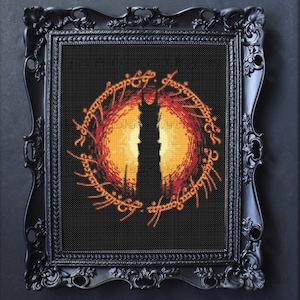 Puede incluir: Obra de arte de punto de cruz del Ojo de Sauron de El Señor de los Anillos, enmarcada en un marco negro ornamentado. La imagen presenta una silueta de torre negra sobre un fondo naranja y rojo ardiente, rodeada de texto dorado.