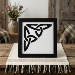 May include: A square black frame containing a white fabric piece with a black cross-stitched triquetra knot design. The embroidery features a geometric Celtic-inspired pattern centred on the fabric, displayed within a simple, clean-lined frame.