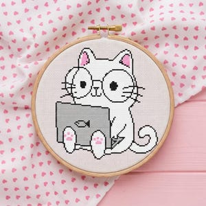 Könnte beinhalten: Eine Kreuzstichstickerei in einem runden Holzrahmen, die eine weiße Katze mit rosa Ohren und Pfoten zeigt, die eine runde Brille trägt und einen grauen Laptop mit einem kleinen Fischsymbol auf dem Deckel benutzt.