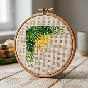 Könnte beinhalten: Ein Stickrahmen mit einem tropischen Design. Das Design zeigt grüne Palmenblätter und eine gelbe Sonne auf beigem Stoff. Der Holzrahmen wird mit einer Messingschraube befestigt.
