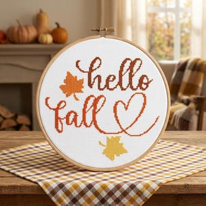May include: A circular wooden embroidery hoop containing white fabric with a cross-stitch design. The text reads hello fall in brown cursive lettering, accompanied by an orange maple leaf, a yellow maple leaf, and a brown heart outline.