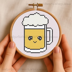 May include: A cross-stitch embroidery of a cartoon beer mug with a smiling face. The mug is yellow with white foam and a black outline, set in a wooden hoop. The background is white.