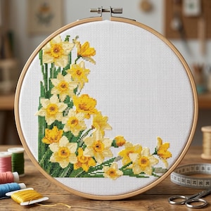 Può includere: Ricamo a punto croce di narcisi gialli e steli verdi in un telaio di legno. Il disegno floreale è nell'angolo del tessuto bianco. Il telaio è fissato con un fermaglio di metallo. Forniture per cucito sullo sfondo.
