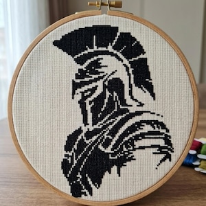 Puede incluir: Un punto de cruz en blanco y negro de un guerrero espartano en un bastidor de bordado de madera. El guerrero está representado de perfil, con casco y armadura. El fondo es de tela beige claro.