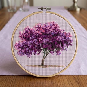 Puede incluir: Un bordado de punto de cruz de un árbol con un dosel púrpura vibrante y un tronco marrón. El bordado está en un aro de madera redondo, sobre un fondo de tela morada clara.