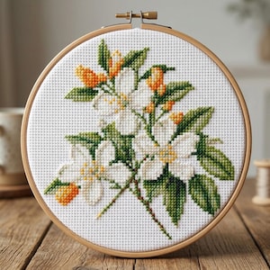 Könnte beinhalten: Kreuzstich-Stickerei mit weißen Blüten, grünen Blättern und orangefarbenen Knospen, eingefasst in einen Holzrahmen. Das florale Design ist auf einem weißen Stoffhintergrund zentriert und zeigt detaillierte Handarbeit. Der Rahmen ist aus hellem Holz gefertigt.