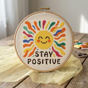 Op de afbeelding: Een borduurring met kruissteek met een lachende gele zon en kleurrijke stralen. De tekst "STAY POSITIVE" is onder de zon geborduurd. De ring staat op een houten oppervlak.