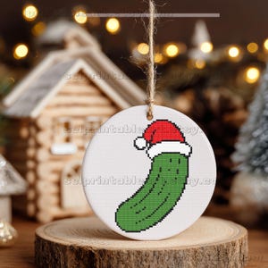 May include: A round, white Christmas ornament featuring a pixelated green pickle wearing a red Santa hat. The ornament hangs from a natural twine string and is displayed on a wooden surface.