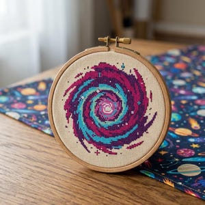 May include: A circular wooden embroidery hoop containing a cross-stitch pattern of a spiral galaxy. The design features vibrant shades of magenta, purple, and teal thread stitched onto a light beige fabric, creating a swirling cosmic effect.