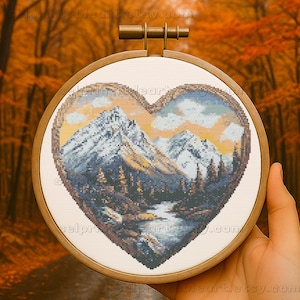 May include: A heart-shaped cross-stitch embroidery depicting a mountain scene with snowy peaks, a river, and trees. The artwork is framed in a round wooden hoop. The background shows a blurred view of an autumn forest.