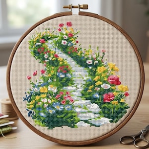 May include: A cross-stitch embroidery of a garden path in a wooden hoop. The path is made of white stones, surrounded by lush greenery and colourful flowers in shades of red, yellow, pink, and blue. The fabric is a light beige.