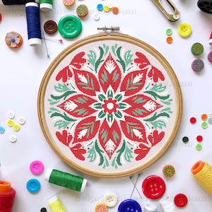 Op de afbeelding: Een voltooide kruissteekborduurwerk in een houten borduurring. Het ontwerp heeft een symmetrisch bloemenpatroon in rood, wit en groen. De achtergrond is een lichtbeige stof. Diverse naaibenodigdheden, waaronder knopen en garen, omringen de ring.