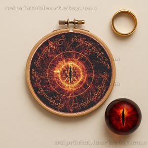 May include: A cross-stitch embroidery in a wooden hoop depicts the Eye of Sauron from Lord of the Rings, with a gold ring and a glass eye-shaped paperweight. The embroidery features a fiery orange and black design with script around the eye.
