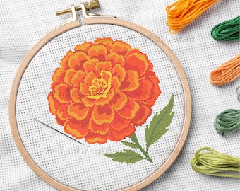 May include: A cross-stitch of an orange marigold flower with green leaves, displayed in a wooden embroidery hoop. The design is accompanied by orange, green, and yellow embroidery threads. The text "selprintableart.etsy.com" is visible.