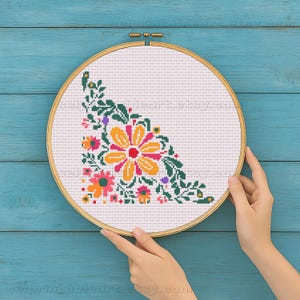 Op de afbeelding: Een kruissteek bloemenontwerp in een houten borduurring. Het ontwerp toont een grote gele bloem met rode en oranje accenten, omringd door groene bladeren en kleinere bloemen in roze, oranje en paars. De achtergrond is wit.