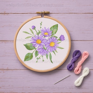 以下が含まれることがあります： 木製の刺繍枠に収められた、黄色い中心と緑の葉を持つ紫色の花のクロスステッチの完成品。紫、ピンク、白の刺繍糸3本と針も写っています。