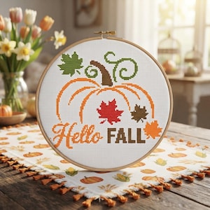 Op de afbeelding: Een ronde houten borduurring met witte stof voorzien van een kruissteekontwerp van een oranje pompoen, groene ranken en kleurrijke herfstbladeren. De tekst Hello FALL is in oranje en bruine draad onder de pompoen geborduurd.