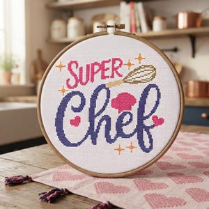 Könnte beinhalten: Ein runder Stickrahmen aus Holz mit weißem Stoff und einem Kreuzstichmotiv. Der Text lautet SUPER in Pink und Chef in Dunkelviolett. Ein goldener Schneebesen, eine pinke Kochmütze und kleine orangefarbene Sterne sind um den Schriftzug gestickt.