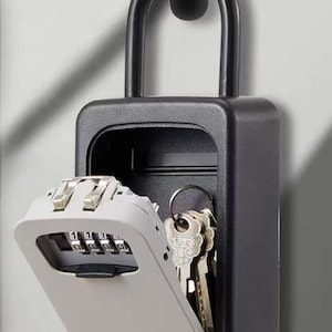 Op de afbeelding: Een zwarte sleutelkluis met een combinatieslot en een scharnierende deur. Het combinatieslot is lichtgrijs met zwarte cijfers. Er zijn verschillende sleutels zichtbaar in de kluis. De kluis hangt aan een zwarte metalen lus.