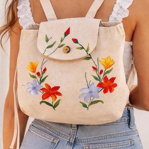 May include: A beige canvas backpack with a flap closure and adjustable straps. The front of the backpack is decorated with hand-painted flowers in shades of red, orange, yellow, and blue. The backpack has a wooden button closure.