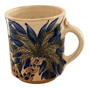 Puede incluir: Taza de cerámica color crema con un diseño de palmera en azul y marrón. La taza tiene un asa redondeada y una forma ligeramente irregular, lo que le da un aspecto hecho a mano. Las hojas de palma y las bayas están pintadas en la taza.