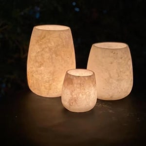 May include: Three glowing, off-white, textured glass candle holders of varying sizes. The largest is oval-shaped, the medium is also oval, and the smallest is a rounded cylinder. The candle holders are lit from within, creating a warm ambiance.