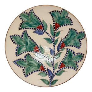 May include: A round ceramic plate with a cream-colored base featuring a hand-painted floral motif. The design includes large green bell-shaped flowers with red centers, blue accents, and dark blue dotted outlines on a central branching stem.