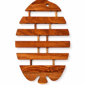 May include: Wooden trivet with a unique fish-like design. The trivet is made of warm-toned wood and features horizontal slats held together by vertical dowels. The top has a rounded shape with a hole, and the bottom has a decorative, curved edge.