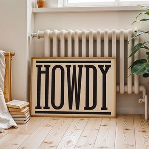 May include: A framed sign with the word "HOWDY" in bold, black letters. The sign has a cream background and a wooden frame. It is propped against a wall near a radiator and a potted plant. The room has wooden flooring.