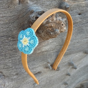Puede incluir: Una diadema marr&oacute;n con un adorno de flor azul y amarilla. La flor est&aacute; hecha de cuentas y tiene un centro blanco.