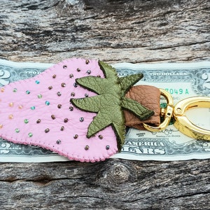 Puede incluir: Un llavero de cuero rosa en forma de fresa con una hoja de cuero verde y un anillo de llave de metal dorado. El llavero est&aacute; sobre un billete de dos euros.