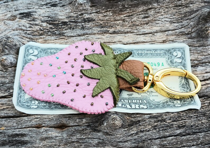 Puede incluir: Un llavero de cuero rosa en forma de fresa con una hoja de cuero verde y un anillo de llave de metal dorado. El llavero est&aacute; sobre un billete de dos euros.