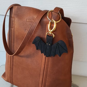 May include: A brown leather tote bag with a black bat-shaped keychain attached to the zipper pull. The keychain is made of fabric and has gold-colored accents.