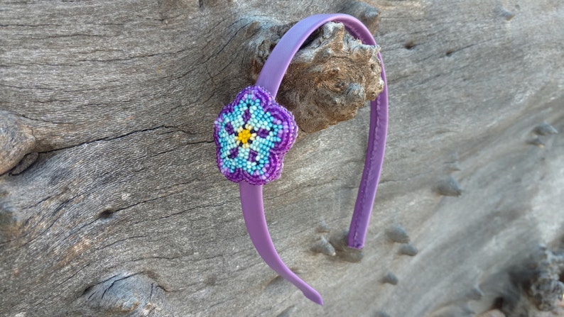 May include: Purple headband with a beaded flower accent. The flower is blue, yellow, and purple.