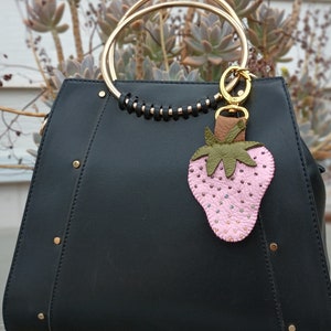 Puede incluir: Un bolso de mano de cuero negro con un asa de anillo dorado y un llavero de fresa de cuero rosa. La fresa est&aacute; adornada con peque&ntilde;as cuentas.