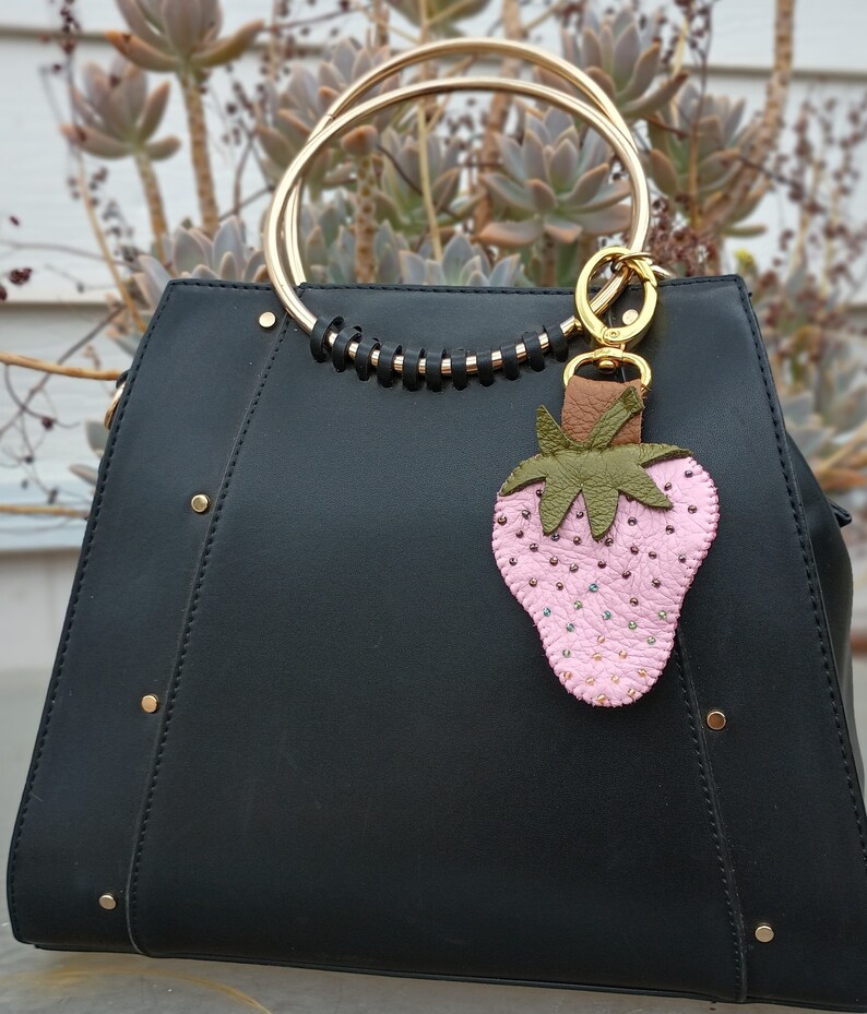 Puede incluir: Un bolso de mano de cuero negro con un asa de anillo dorado y un llavero de fresa de cuero rosa. La fresa est&aacute; adornada con peque&ntilde;as cuentas.